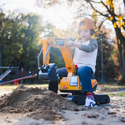 A young child in a cap uses the TP Toys Falk Power Builder Digger with Opening Seat to scoop sand outdoors, with trees and playground equipment visible in the background.