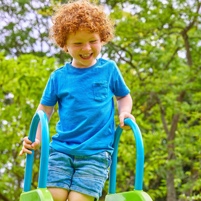 A young child with curly red hair, in a blue shirt and denim shorts, smiles as they climb a ladder beside the TP Toys TP Small to Tall Growable Slide, enjoying this fun and adjustable outdoor playset.
