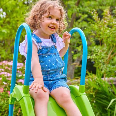 A young child sits atop the TP Toys TP Small to Tall Growable Slide outdoors, wearing a white shirt and denim overalls, surrounded by greenery and flowers.