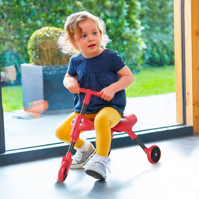 A young child in a navy shirt and yellow pants rides the Scuttlebug Beetle Foldable Ride On Trike by Scuttlebug indoors near a large window.