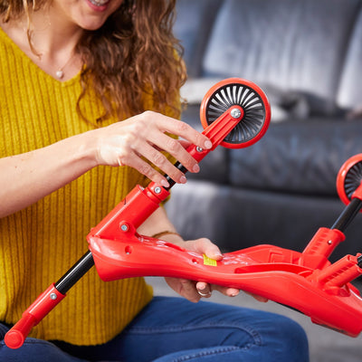 A person in a yellow sweater assembles a Scuttlebug Beetle Foldable Ride On Trike by Scuttlebug, attaching a wheel while sitting indoors.