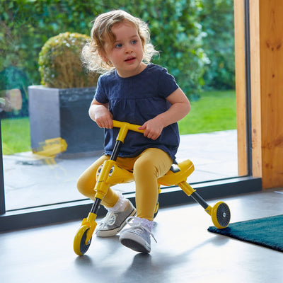 A young child in a blue shirt and yellow pants sits indoors near a large window on a TP Toys Scuttlebug Bumble Foldable Ride On Trike.