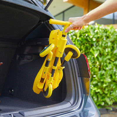 A hand places a folded yellow TP Toys Scuttlebug Bumble Foldable Ride On Trike into the open trunk of a car, with greenery in the background.