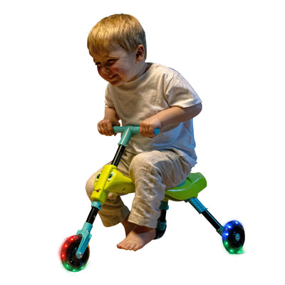 A young child in light-colored clothes rides a TP Toys Scuttlebug Firefly, a small yellow and teal three-wheeled trike with motion-powered light-up wheels and illuminated red and blue training wheels against a white background.