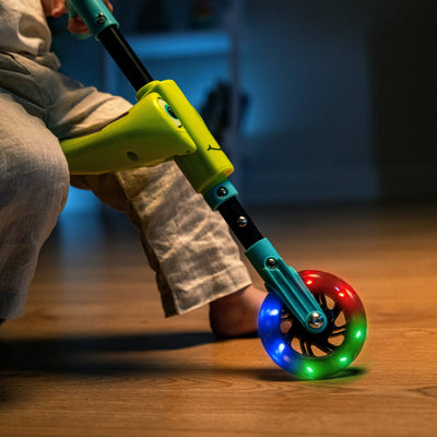 A child sits indoors on a TP Toys Scuttlebug Firefly, a three-wheeled ride-on scooter with a blue handle and motion-powered wheels that light up red, green, and blue.