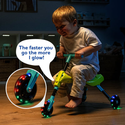 A young child rides a TP Toys Scuttlebug Firefly indoors, its motion-powered LED wheels glowing brighter the faster they go. A speech bubble says, The faster you go the more I glow! A close-up shows off the light-up wheels.