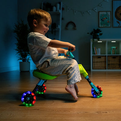 A young child rides the TP Toys Scuttlebug Firefly, a three-wheeled ride-on with motion-powered light-up wheels, indoors on a wooden floor, surrounded by home decor.