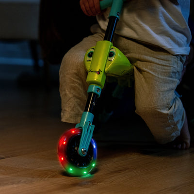 A child rides the TP Toys Scuttlebug Firefly, a three-wheeled indoor scooter with a firefly design and motion-powered wheels that light up in red, blue, and green.