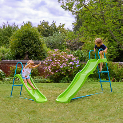 Two children play on the TP Toys TP Small to Tall Growable Slide in a grassy backyard, surrounded by trees and flowering bushes beneath a cloudy sky.