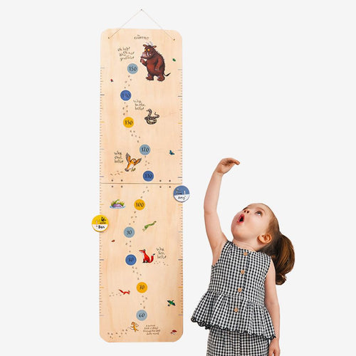 A young girl stands beside The Gruffalo Wooden Height Chart - FSC® certified by TP Toys, decorated with animal illustrations, reaching up to measure her height.