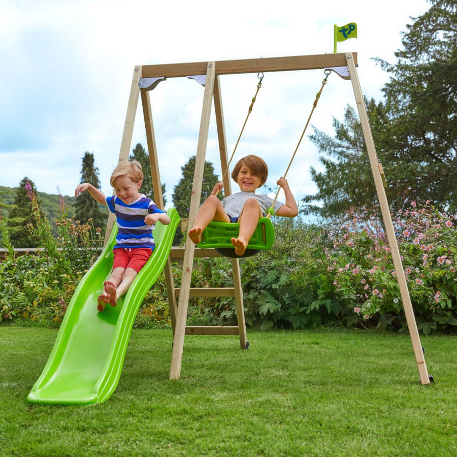 Two children enjoy the TP Toys TP Forest Multiplay Single Wooden Swing Set with Roped Rapide Seat & Slide (FSC® certified)—one swings while the other slides—outdoor fun on a grassy lawn with trees and flowers in the background.