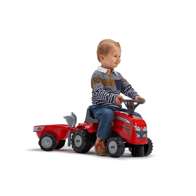 A young child wearing jeans and a striped sweater sits on the TP Toys Falk Baby Massey Ferguson Ride-on Tractor with Trailer, Rake & Shovel, made from recycled plastic. The toddler outdoor toy is set against a plain white background.