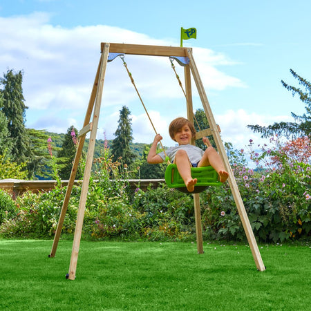 A child sits and smiles on the TP Toys Forest Wooden Single Swing Set with Roped Rapide Seat (FSC® certified) in a grassy, flower-filled garden surrounded by trees.