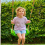 A young child with curly hair smiles while sitting on a green swing attached to the TP Toys Single Wooden Swing Arm for Buttercup Cottage Playhouse (FSC® certified), surrounded by leafy bushes.