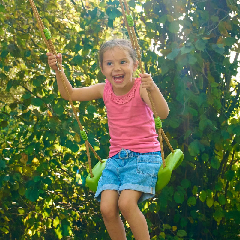 A young girl in a pink top and denim shorts smiles as she swings outdoors on the TP Toys Double Wooden Swing Arm for Buttercup Cottage Playhouse (FSC® certified), surrounded by greenery.