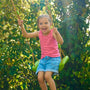 A young girl in a pink top and denim shorts smiles as she swings outdoors on the TP Toys Double Wooden Swing Arm for Buttercup Cottage Playhouse (FSC® certified), surrounded by greenery.