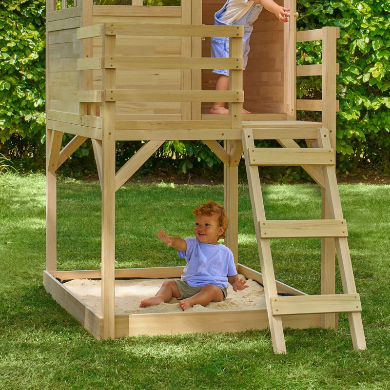 A young child plays in a sandbox under the TP Toys Wooden Deck for Buttercup Cottage Playhouse, made from FSC® certified wood, while another stands above. They are in a grassy backyard bordered by lush green bushes.