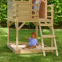 A young child plays in a sandbox under the TP Toys Wooden Deck for Buttercup Cottage Playhouse, made from FSC® certified wood, while another stands above. They are in a grassy backyard bordered by lush green bushes.