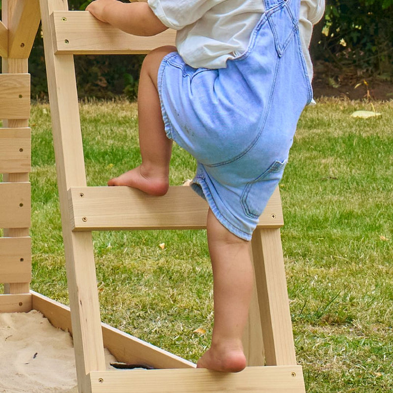 A young child in a light blue overall dress climbs the FSC® certified Wooden Deck for Buttercup Cottage Playhouse by TP Toys outdoors, with grass and trees in the background.