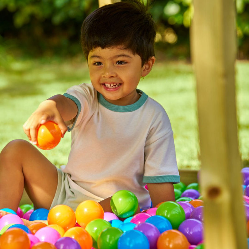 A young child smiles in a ball pit outdoors at Buttercup Playhouse, holding an orange plastic ball on the TP Toys Wooden Deck for Buttercup Cottage Playhouse, made from FSC® certified wood.