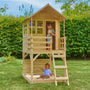 Two children play in the TP Toys Buttercup Cottage Wooden Playhouse with Verandah and Deck—FSC® certified—in a grassy backyard. One is inside the cottage while the other enjoys the sandbox on the deck below.