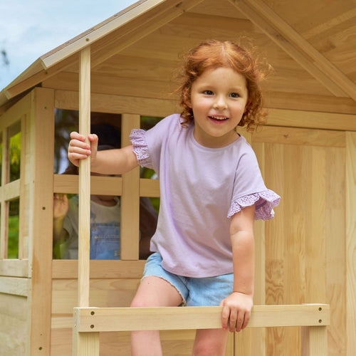 A red-haired child in a lavender shirt and denim shorts smiles on the porch of the TP Toys Buttercup Cottage Wooden Playhouse with Verandah, 6ft Wavy Slide, Swing Arm and Deck—FSC® certified.