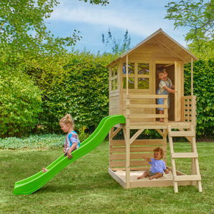 Three children play on the TP Toys Buttercup Cottage Wooden Playhouse with Verandah, 6ft Wavy Slide, and Deck—made from FSC® certified wood—in a grassy backyard surrounded by trees and bushes.