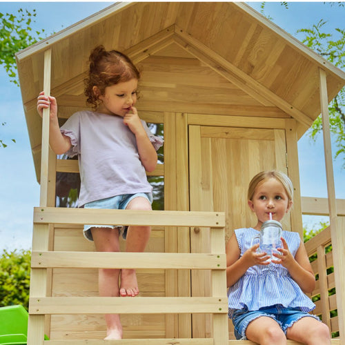 Two young girls enjoy the TP Toys Buttercup Cottage Wooden Playhouse with Verandah, 6ft Wavy Slide, Double Swing Arm and Deck—one sits barefoot on the steps while the other drinks from a clear cup with a straw.