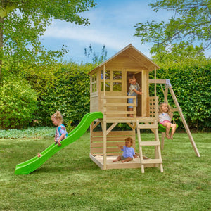 Four young children play on the TP Toys Buttercup Cottage Playhouse with a Single Wooden Swing Arm—made from FSC® certified wood—featuring a green slide and ladder, all set on a grassy lawn with trees and bushes nearby.