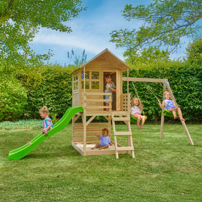Five children play on the TP Toys Buttercup Cottage Wooden Playhouse with Verandah, 6ft wavy slide, double swing arm and deck—made from FSC® certified wood—in a grassy backyard surrounded by trees and bushes.