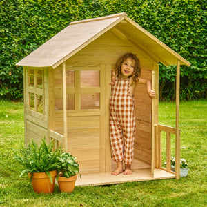 A young child stands smiling in the doorway of the TP Toys Buttercup Cottage Wooden Playhouse with Floor and Verandah—crafted from FSC® certified wood—in a garden, with potted plants nearby.