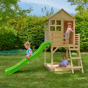 Three young children play on the TP Toys Buttercup Cottage Wooden Playhouse with Deck & 6ft Wavy Slide—FSC® certified—in a grassy backyard bordered by bushes and trees.