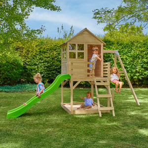 Four children enjoy the Buttercup Cottage Wooden Playhouse by TP Toys, featuring a deck, swing arm, and 6ft wavy slide in a grassy backyard with bushes and trees.