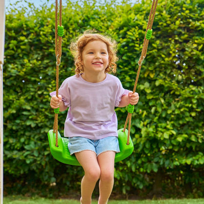 A young child with curly hair smiles on a green swing, part of the TP Toys Buttercup Cottage Wooden Playhouse with Deck, Double Swing Arm & 6ft Wavy Slide—FSC® certified—with green bushes in the background.