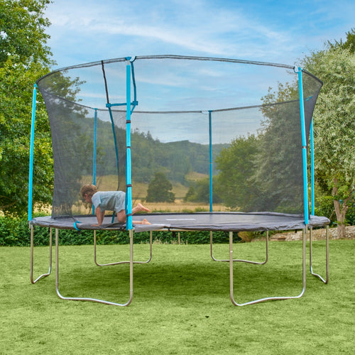 A child kneels on a TP Toys TP Up 14ft Trampoline with a safety net in the backyard, surrounded by green grass and trees under a blue sky.