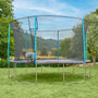 A child kneels on a TP Toys TP Up 14ft Trampoline with a safety net in the backyard, surrounded by green grass and trees under a blue sky.