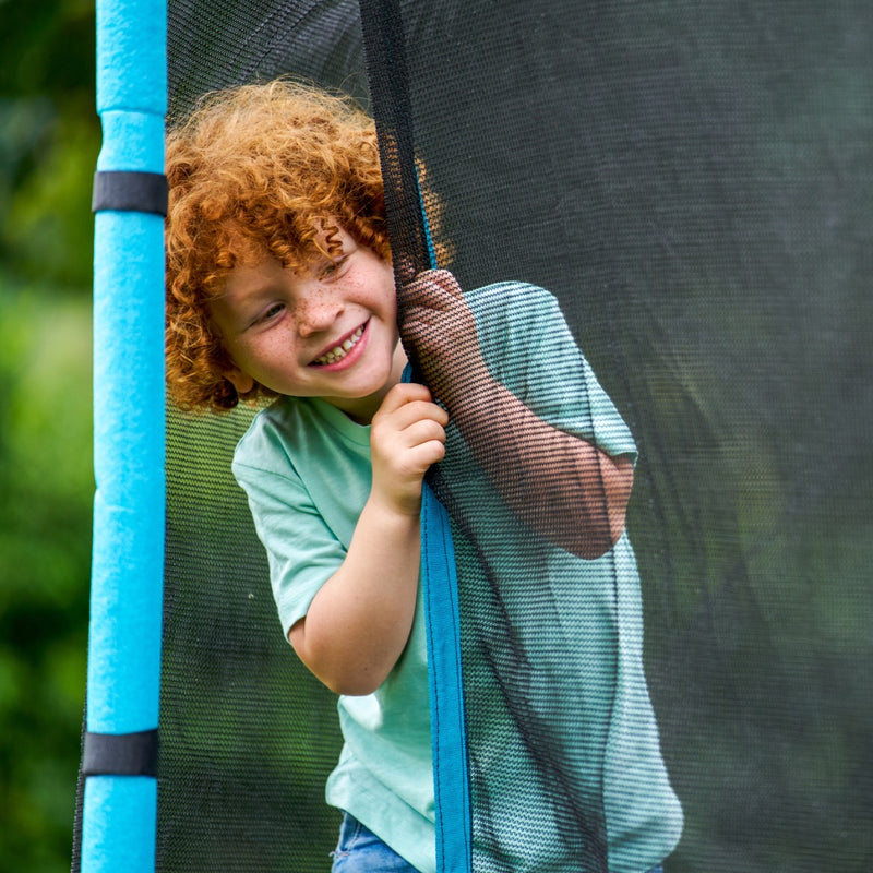 A young child with curly red hair and a green shirt smiles while peeking out from behind the safety net of a TP Toys TP Up 14ft Trampoline, highlighting the fun and excitement it brings to kids.