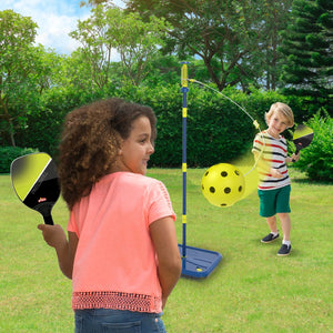 Two children play with the TP Toys Pickleball All Surface Swingball on a grassy lawn. An adjustable pole from the set stands between them, surrounded by trees and bushes in the background.