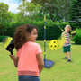 Two children play with the TP Toys Pickleball All Surface Swingball on a grassy lawn. An adjustable pole from the set stands between them, surrounded by trees and bushes in the background.