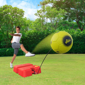 A boy perfects his skills with the TP Toys Swingball Reflex Soccer, kicking a yellow ball attached by a cord to an All Surface base in a grassy outdoor area with trees behind him.