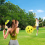 Two people play pickleball on a grassy field using the TP Toys Pickleball Bat and Ball Set; one gets ready to hit the lightweight ball while the other jumps to return it. Trees and a blue sky can be seen in the background.