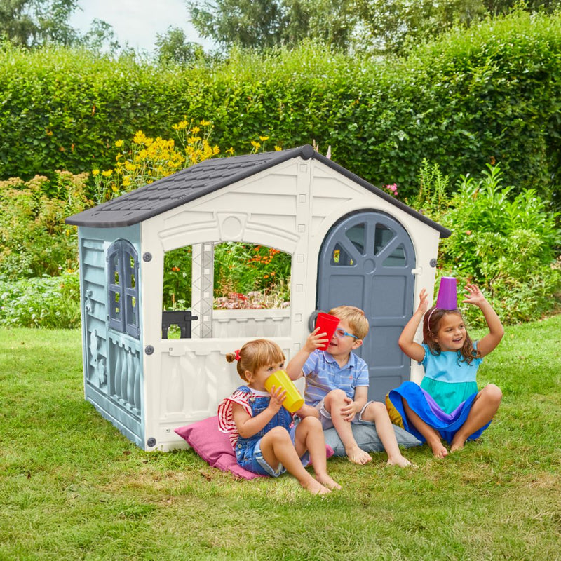 Three children sit on cushions in a garden outside the TP Toys Palplay Plastic Misty Sky Playhouse of Fun, each holding a colorful cup to their mouth or head.