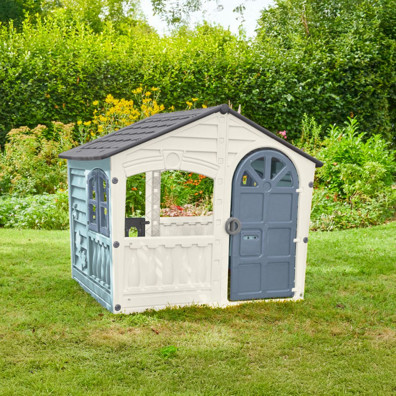 The TP Toys Palplay Plastic Misty Sky Playhouse of Fun, a UV-resistant white and gray playhouse with door and windows, is shown on garden grass amid green plants and yellow flowers in the background.