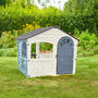 The TP Toys Palplay Plastic Misty Sky Playhouse of Fun, a UV-resistant white and gray playhouse with door and windows, is shown on garden grass amid green plants and yellow flowers in the background.