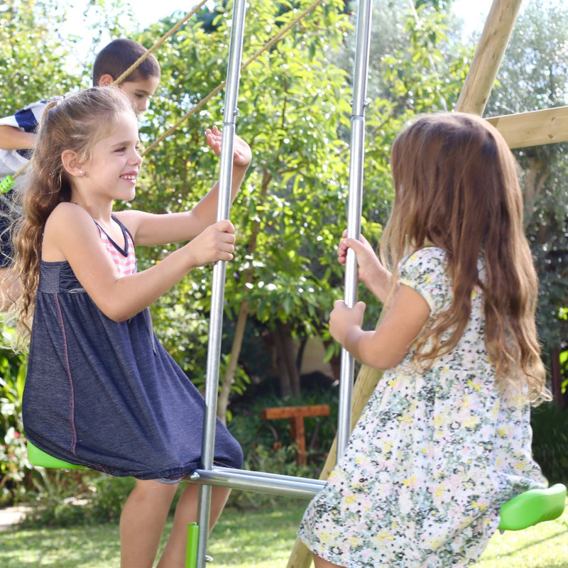 Two young girls smile and face each other on the TP Toys Knightswood Double Wooden Swing Set With Glide Ride, while a boy stands in the background outdoors on a sunny day.