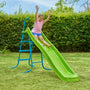 A girl in a pink shirt and blue shorts enjoys the TP Toys TP 8ft (2.2m) Ripple Wavy Slide & Stepset, sliding down with raised hands in a grassy yard with a hedge in the background.