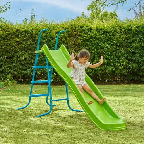 A young child enjoys outdoor play, sliding down the TP Toys 8ft (2.2m) Ripple Wavy Slide & Stepset in a grassy backyard surrounded by bushes and trees.
