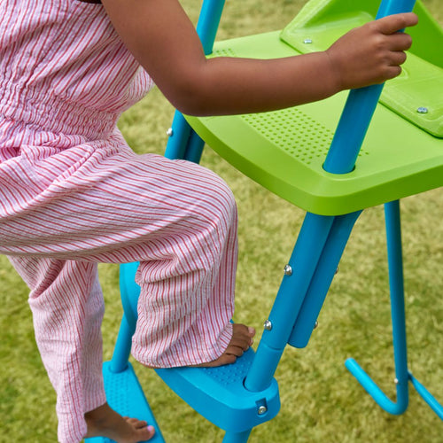 A child in pink and white stripes climbs the blue and green steps of the TP Toys TP 8ft (2.2m) Ripple Wavy Slide & Stepset, making for a fun kids' garden slide adventure on the grass.