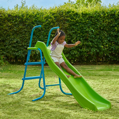 A young child slides down the TP Toys TP 6ft (1.8m) Wavy Slide & Stepset in a grassy backyard garden playground with a hedge in the background.