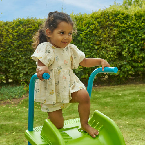 A young girl stands atop the TP Toys 6ft (1.8m) Wavy Slide & Stepset, holding blue handles, with grass and hedges in the background—perfect for any garden playground.